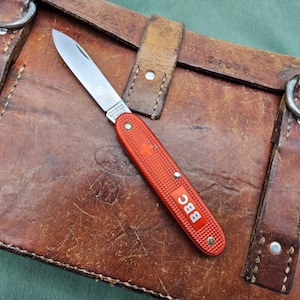 May include: A folding pocket knife with a single silver-colored steel blade and a textured orange handle. The handle features a small white Swiss cross emblem and the white text "BBC" printed on the side. The knife is in an open position.