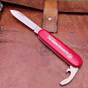 May include: A red pocket knife with a silver blade and bottle opener. The knife has the text "SONNENBRAU REBSTEIN" printed on the side. The knife is resting on a brown leather surface.