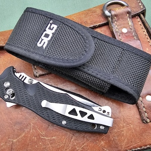May include: A black SOG folding knife with a silver pocket clip and a black nylon sheath. The knife is open, revealing the blade. The sheath has the SOG logo in white. The knife and sheath are on a brown leather surface.