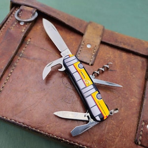 May include: A multi-tool knife with a yellow and gray design, featuring various tools like a blade, bottle opener, and corkscrew. The knife rests on a brown leather case with straps and metal rings.