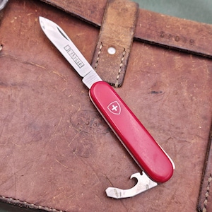 May include: A red Swiss Army knife with a silver blade and a bottle opener. The knife is on a brown leather surface.