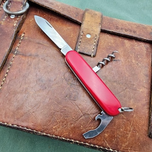 May include: A red pocket knife with a corkscrew, bottle opener, and blade. The knife is lying on a brown leather surface.