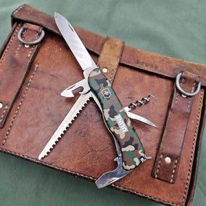 Könnte beinhalten: Ein Multitool-Messer mit Camouflage-Griff, mit Klinge, Säge, Flaschenöffner und Korkenzieher, liegt auf einem braunen Lederetui mit Riemen und Metallringen. Auf dem Messer steht das Wort "ULINE".