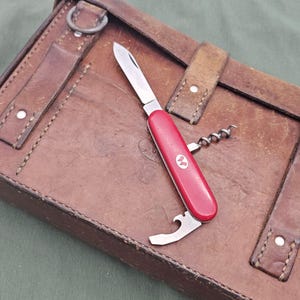 May include: A red multi-tool knife with a corkscrew and other tools, resting on a brown leather case. The knife has a silver blade and a white emblem on the handle. The case has straps and metal rings.