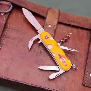 May include: A yellow Swiss Army knife with white polka dots and the word "Switzerland" in red. The knife has multiple tools, including a blade, bottle opener, and corkscrew. It rests on a brown leather bag.