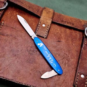 Könnte beinhalten: Ein blaues Taschenmesser mit silberner Klinge und einer kleineren Sekundärklinge. Das Messer hat einen blauen Griff mit weißem Text und einem Sonnenmuster. Es liegt auf einer braunen Lederoberfläche.