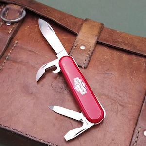 Op de afbeelding: Een rood en zilver multi-tool mes met de merknaam "Hürlimann" op de zijkant. Het mes is open en toont een mes, flesopener en andere gereedschappen. Het ligt op een bruin leren oppervlak.