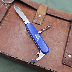 May include: A blue multi-tool knife with a corkscrew, bottle opener, and blade, resting on a brown leather surface. The knife has the brand name "Herberts" printed on the handle.