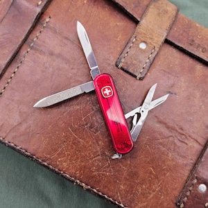 May include: A red Swiss Army knife with multiple tools, including a blade, nail file, and scissors, is open on a brown leather surface. The knife features a white cross on a red background. The leather has visible wear and tear.