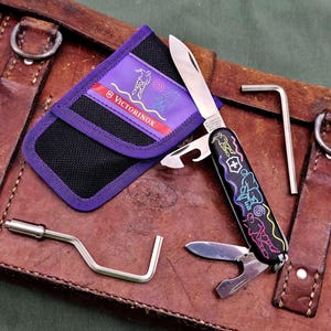 Könnte beinhalten: Ein schwarzes Multitool-Messer mit einem farbenfrohen Design mit Sportfiguren, einer Klinge und einem Flaschenöffner. Das Messer liegt neben einem lila-schwarzen Etui mit dem Victorinox-Logo und zwei Metallwerkzeugen, alles auf einer braunen Lederoberfläche.