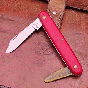 Pode incluir: Uma faca de bolso vermelha com uma lâmina prateada e uma ferramenta de cor latão. A faca está aberta, revelando a lâmina. A pega é de cor vermelha vibrante. A faca está pousada numa superfície de couro castanho.