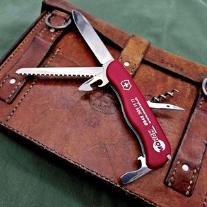 Könnte beinhalten: Ein rotes Multifunktions-Taschenmesser mit silberner Klinge, Säge, Flaschenöffner und Korkenzieher. Der Griff zeigt den weißen Schriftzug MOIKAH, eine Telefonnummer und eine Webadresse sowie ein kleines weißes Schweizerkreuz-Emblem an der Seite.