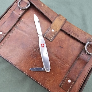 Puede incluir: Una navaja suiza plateada con un emblema de cruz roja, hoja abierta y una hoja más pequeña, sobre un estuche de cuero marrón con correas y anillos metálicos. El estuche está desgastado.