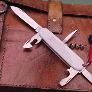 Könnte beinhalten: Ein weißes Schweizer Taschenmesser mit einem roten Band. Das Messer verfügt über eine Vielzahl von Werkzeugen, darunter eine Klinge, einen Korkenzieher, einen Dosenöffner und einen Schraubendreher.