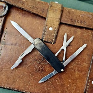 Peut inclure: Un couteau de poche multifonction doté d'un manche sombre et texturé, avec plusieurs outils en acier inoxydable déployés, dont une lame principale, un outil dentelé, une paire de petits ciseaux et un tournevis plat à la finition polie.