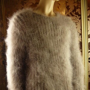 Peut inclure: Un pull à manches longues et col rond de couleur taupe clair ou brun grisâtre. Le vêtement présente une maille très texturée, duveteuse et brossée, avec un motif côtelé vertical visible sur tout le corps et les manches.