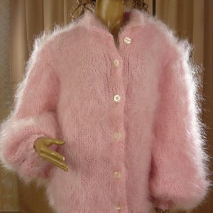 Può includere: Un cardigan in maglia soffice rosa chiaro a maniche lunghe, caratterizzato da un colletto alto e una chiusura frontale con cinque bottoni bianchi rotondi. Il capo presenta una vestibilità ampia con maniche voluminose e una consistenza morbida.