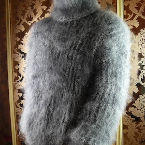 Peut inclure: Un pull à manches longues gris, duveteux et à col haut, doté d'un tricot épais et texturé. Le vêtement présente un col roulé haut et proéminent ainsi qu'un aspect lâche et hirsute créé par les longues fibres douces du fil sur toute la pièce.
