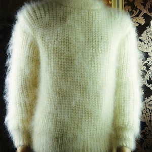 Peut inclure: Un pull à manches longues de couleur crème doté d'une maille épaisse, duveteuse et texturée. Le vêtement présente une encolure ronde, des poignets côtelés aux manches et un ourlet côtelé, offrant un aspect doux, volumineux et poilu.