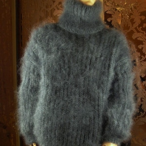 Peut inclure: Un pull en tricot duveteux gris foncé à manches longues avec un col roulé haut. Le vêtement présente un aspect épais et texturé avec des motifs de côtes verticales visibles le long des manches et du corps, porté sur un mannequin.