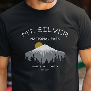 Może przedstawiać: Czarny T-shirt z białym nadrukiem graficznym przedstawiającym pasmo górskie z żółtym słońcem. Tekst "MT. SILVER NATIONAL PARK" znajduje się nad górą, a "ROUTE 28 - JOHTO EST 1997" pod górą.