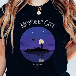 May include: Black t-shirt featuring a circular graphic of a city silhouette at night, with a full moon and stars. The text "MOSSSDEEP CITY" arches above the image, with "HOENN EST 2002" below. The design is reflected in the water.