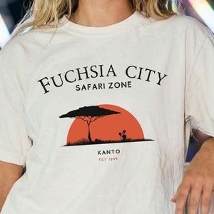 Pode incluir: Uma t-shirt de manga curta na cor creme com uma estampa gráfica contendo o texto "FUCHSIA CITY SAFARI ZONE" acima de uma silhueta de sol laranja, uma árvore preta e o texto "KANTO EST 1996" abaixo do gráfico na frente da peça.