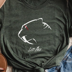 Puede incluir: Una camiseta verde con un gráfico blanco de un tigre dientes de sable con una barra roja sobre el ojo. El texto "Lost Age" está impreso debajo del gráfico.