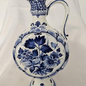 Könnte beinhalten: Ein weißer Keramikkrug mit komplexen blauen Blumenmustern und Blattmotiven. Das Gefäß hat einen runden Körper, einen schlanken Hals mit einem ausgestellten Ausguss, einen geschwungenen Henkel und einen runden Sockelfuß, verziert mit blauen Motiven.