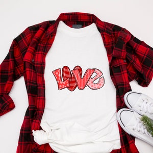 以下が含まれることがあります： 赤いバブルスタイルのフォントで「LOVE」という言葉がプリントされた白いTシャツ。水玉模様とストライプがアクセントになっています。赤と黒のチェック柄フランネルシャツ、白いスニーカー、小さな常緑樹の小枝と組み合わせています。