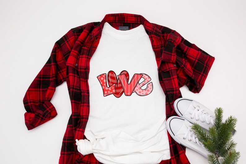以下が含まれることがあります： 赤いバブルスタイルのフォントで「LOVE」という言葉がプリントされた白いTシャツ。水玉模様とストライプがアクセントになっています。赤と黒のチェック柄フランネルシャツ、白いスニーカー、小さな常緑樹の小枝と組み合わせています。