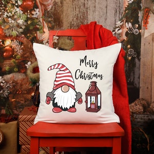 May include: A square, off-white Christmas pillow featuring a gnome illustration with a red and white striped hat, a white beard, and a lantern. The words "Merry Christmas" are written in black script.