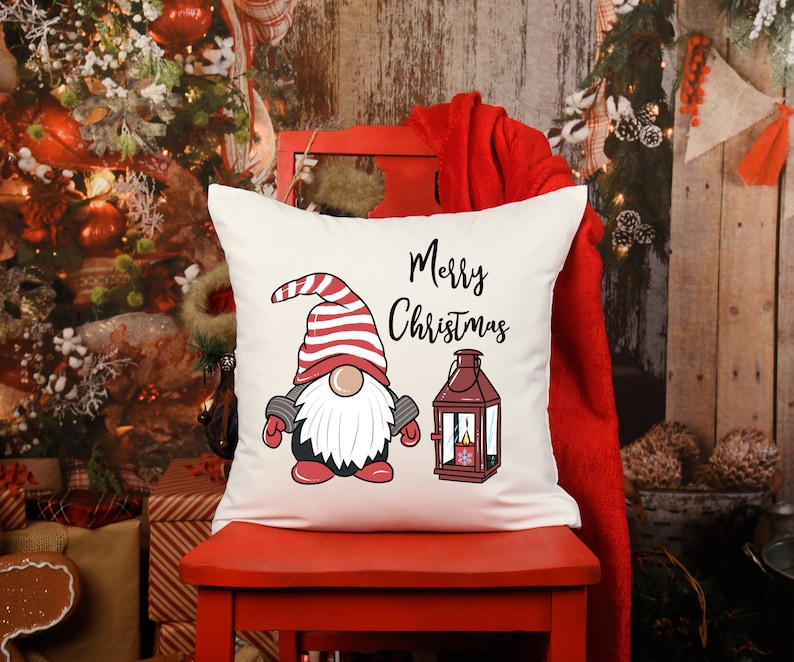 May include: A square, off-white Christmas pillow featuring a gnome illustration with a red and white striped hat, a white beard, and a lantern. The words "Merry Christmas" are written in black script.