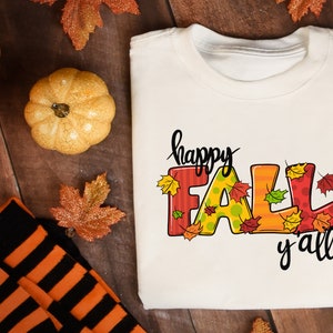May include: A white t-shirt with the words "happy FALL y'all" in a colorful, fall-themed design. The word "FALL" is in large, decorated letters with autumn leaves. The shirt is folded on a wooden surface, with pumpkins and fall leaves.