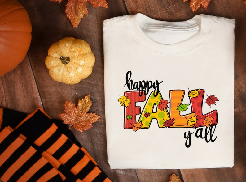 May include: A white t-shirt with the words "happy FALL y'all" in a colorful, fall-themed design. The word "FALL" is in large, decorated letters with autumn leaves. The shirt is folded on a wooden surface, with pumpkins and fall leaves.