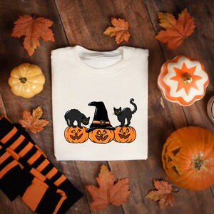 May include: Cream-colored sweatshirt featuring a Halloween design with three orange jack-o'-lanterns, black cats, and a witch's hat. The design is surrounded by fall leaves, pumpkins, and a pair of black and white sneakers.