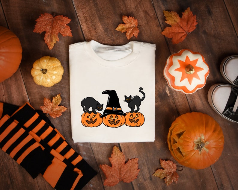 May include: Cream-colored sweatshirt featuring a Halloween design with three orange jack-o'-lanterns, black cats, and a witch's hat. The design is surrounded by fall leaves, pumpkins, and a pair of black and white sneakers.