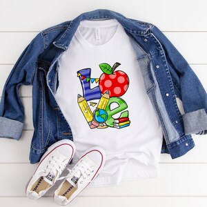 May include: White t-shirt with a colorful "LOVE" graphic, featuring an apple, pencils, globe, and books. A denim jacket and white sneakers are laid out on a white wooden surface, creating a casual, back-to-school theme.