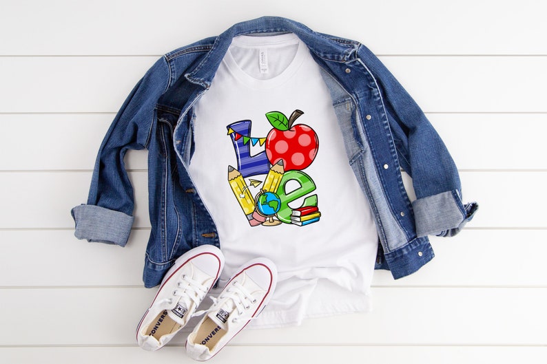 May include: White t-shirt with a colorful "LOVE" graphic, featuring an apple, pencils, globe, and books. A denim jacket and white sneakers are laid out on a white wooden surface, creating a casual, back-to-school theme.