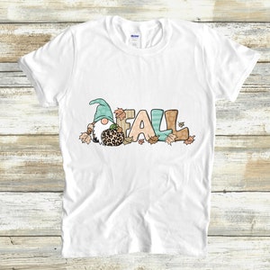 May include: White t-shirt featuring a whimsical design with the word "FALL" in colorful letters. A gnome with a teal hat and a leopard-print pumpkin is part of the design, surrounded by autumn leaves. A fun fall-themed graphic tee.