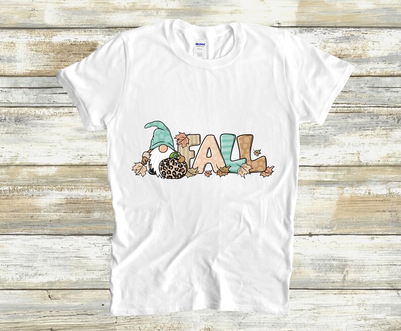 May include: White t-shirt featuring a whimsical design with the word "FALL" in colorful letters. A gnome with a teal hat and a leopard-print pumpkin is part of the design, surrounded by autumn leaves. A fun fall-themed graphic tee.
