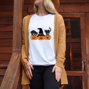 May include: White t-shirt featuring a Halloween design. The graphic includes three orange pumpkins with carved faces, a witch's hat, and two black cats. The shirt is being worn by a person with a tan cardigan.