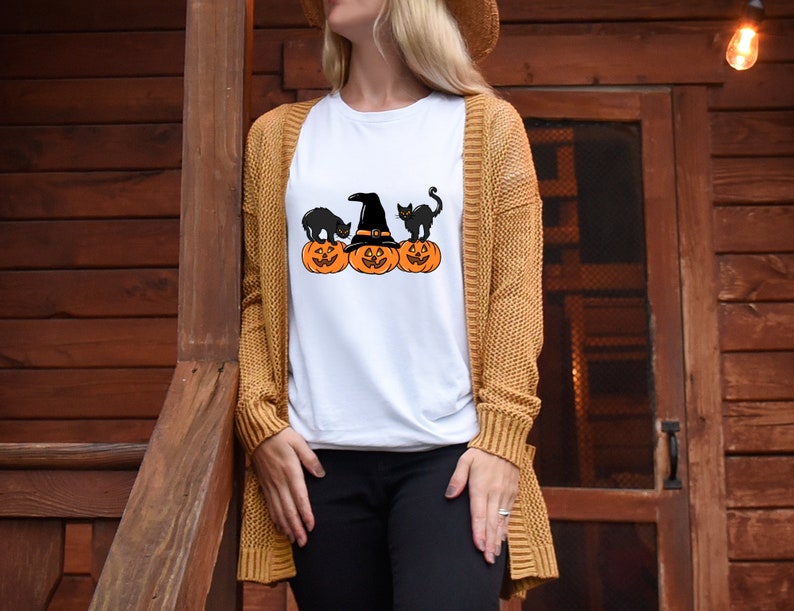 May include: White t-shirt featuring a Halloween design. The graphic includes three orange pumpkins with carved faces, a witch's hat, and two black cats. The shirt is being worn by a person with a tan cardigan.