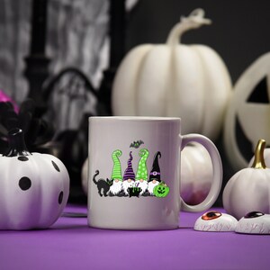 May include: A light gray ceramic mug featuring a Halloween gnome design with green, purple, and black hats, a black cat, and a green pumpkin. White pumpkins and decorative eyeballs are in the background.