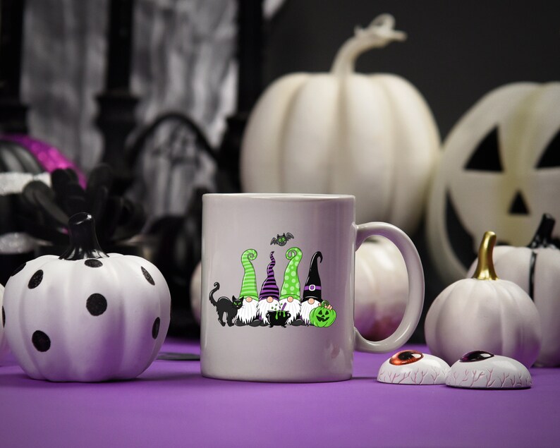 May include: A light gray ceramic mug featuring a Halloween gnome design with green, purple, and black hats, a black cat, and a green pumpkin. White pumpkins and decorative eyeballs are in the background.