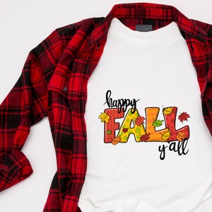 May include: White t-shirt with the words "happy FALL y'all" in black script and colorful letters. The letters are decorated with fall leaves. A red and black plaid shirt is draped over the t-shirt.