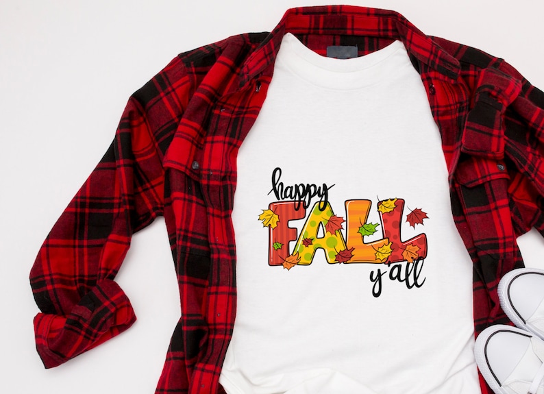 May include: White t-shirt with the words "happy FALL y'all" in black script and colorful letters. The letters are decorated with fall leaves. A red and black plaid shirt is draped over the t-shirt.