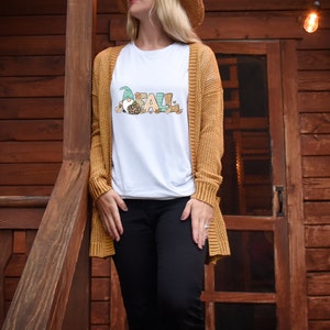 May include: White t-shirt with the word "FALL" in colorful letters and a gnome graphic. The gnome wears a green hat and has a leopard print shirt. The person is wearing a mustard yellow cardigan and black pants.