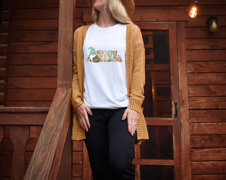 May include: White t-shirt with the word "FALL" in colorful letters and a gnome graphic. The gnome wears a green hat and has a leopard print shirt. The person is wearing a mustard yellow cardigan and black pants.