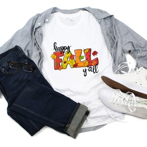 May include: A white t-shirt with the words "happy FALL y'all" in colorful letters and fall leaf accents. The shirt is paired with blue jeans, a gray button-down shirt, and white sneakers.